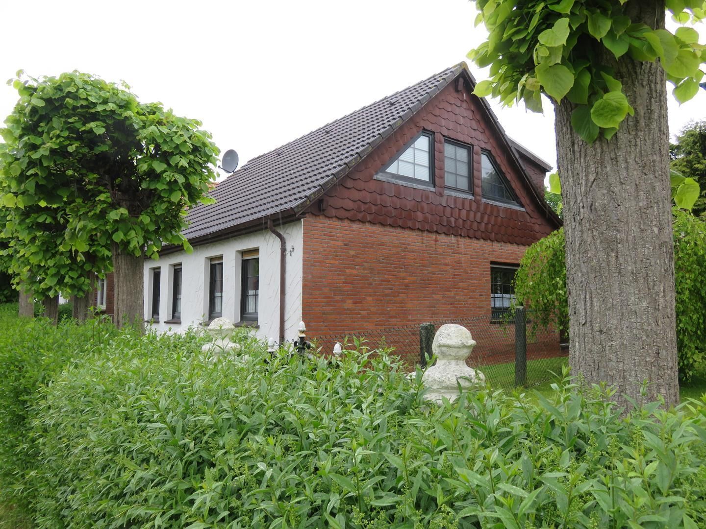 A house with a sloped roof, white exterior walls, and a brick section. There is a tree with green leaves in the foreground, and a fence with a white decorative element.