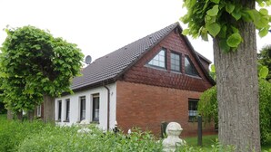 A house with a sloped roof, white exterior walls, and a brick section. There is a tree with green leaves in the foreground, and a fence with a white decorative element.