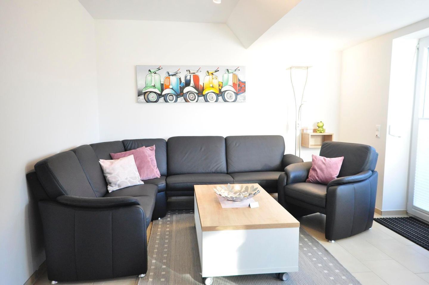 Ein modernes Wohnzimmer mit einem schwarzen Ledersofa, einem weißen Couchtisch und einem farbenfrohen Kunstwerk mit Oldtimern an der Wand.
