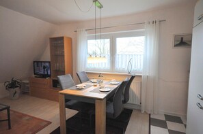 A dining area with a table set for two, a television on a wooden cabinet, and a window with curtains.