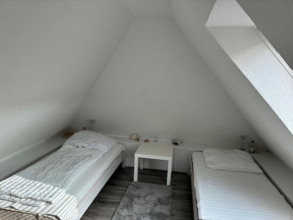 A small, white, attic-style room with two single beds, a small table, and a window. - Vacation Apartment, Fischerstraße 1 (Wittmund)