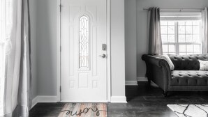 A room with a white door featuring a decorative glass panel, a dark-colored tufted sofa, and a large window with curtains. - Central Charm: 5min to Broadway & DT, Free Coffee! (Nashville)