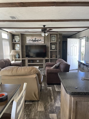A compact living space with a flat-screen TV, two sofas, a dining table, and a kitchen area with a countertop.