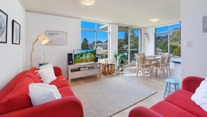 A modern living room with a red sofa, a TV, and a dining area with a table and chairs. - Seaview 7 - Prime Position Beachside Holiday (AVOCA BEACH)