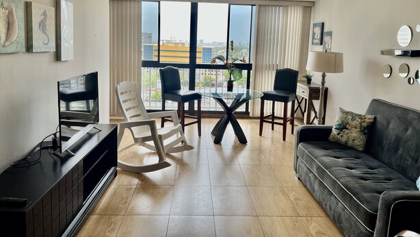 Una sala de estar moderna con una mesa de comedor de cristal, dos sillas, un sofá, un televisor y un ventilador de techo.