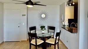 Un comedor con una mesa de vidrio, cuatro sillas, un ventilador de techo, un espejo en la pared y una pequeña planta en maceta.