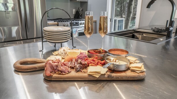 Un plan de travail de cuisine en acier inoxydable, agrémenté d’une planche à découper pour la charcuterie, de deux coupes de champagne et d’une cuisinière en arrière-plan.