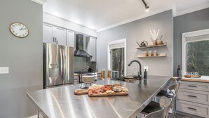 Une cuisine moderne dotée d’appareils électroménagers en acier inoxydable, d’un grand îlot central avec une planche à découper pour la charcuterie, et d’une fenêtre offrant une vue sur l’extérieur.