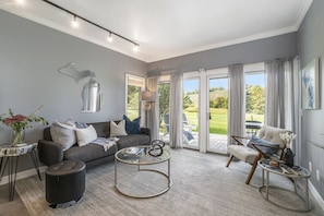 Un salon moderne avec un canapé gris, une table basse ronde et un fauteuil confortable. Une grande fenêtre avec des rideaux, une petite table d’appoint avec un vase et une vue sur un paysage verdoyant complètent l’ensemble.