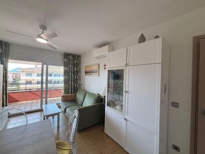 A compact living space with a dining area, a sofa, a glass-fronted refrigerator, and a balcony with a view of buildings.