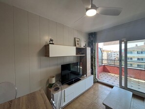 Un salon moderne doté d’un téléviseur à écran plat, de murs recouverts de panneaux en bois, d’un ventilateur de plafond et d’un balcon offrant une vue sur les autres immeubles.