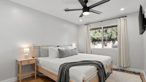 Un dormitorio con cama, mesita de noche, lámpara, ventilador de techo, ventana con cortinas y vistas al exterior.