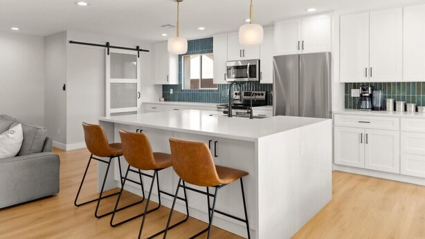 Una cocina moderna con una isla central, taburetes de bar, armarios blancos y electrodomésticos de acero inoxidable.