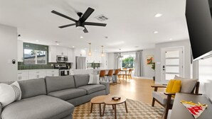 Una sala de estar moderna con un sofá seccional gris, una zona de comedor con sillas de madera y una cocina al fondo.