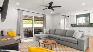 Una sala de estar moderna con un sofá modular gris, una mesa de centro de madera y un cojín amarillo. Hay una zona de comedor con sillas de madera y una mesa, y una cocina con armarios blancos y un salpicadero verde.
