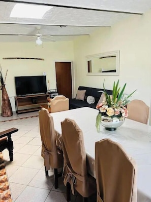 Un comedor con una mesa blanca, sillas color beige, un televisor, un sofá y un jarrón decorativo con flores.