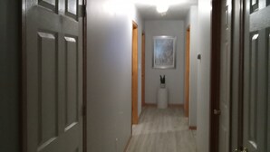 Un pasillo con puertas de madera, una pared blanca con un cuadro enmarcado y un pedestal blanco con una planta.