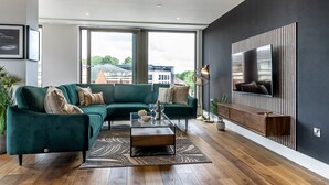 A modern living room with a green sectional sofa, a glass coffee table, and a large flat-screen TV mounted on a dark wall. - The Lux @ Ryedale House -  a premium stay that sleeps 4 guests in 2 bedrooms (York)
