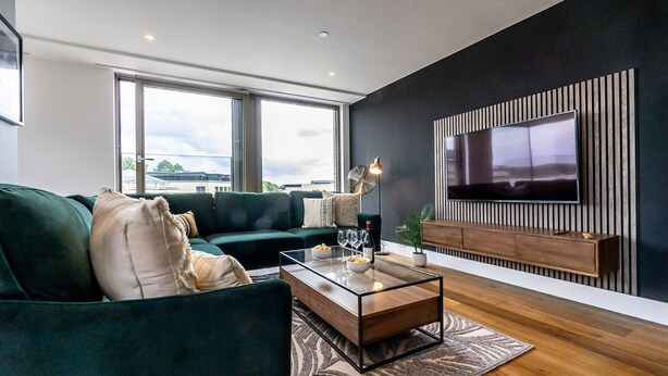 A modern living room with a green sofa, a glass coffee table, and a flat-screen TV mounted on a dark wall. - The Lux @ Ryedale House -  a premium stay that sleeps 4 guests in 2 bedrooms (York)