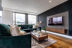 A modern living room with a green sofa, a glass coffee table, and a flat-screen TV mounted on a dark wall.