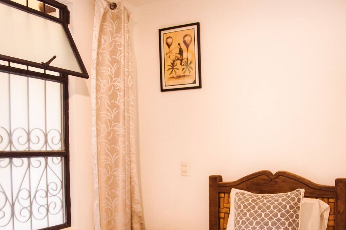 Una cama bien hecha con una toalla doblada, un cabecero de madera, una mesita de noche con un libro, una ventana con barrotes y un cuadro en la pared.