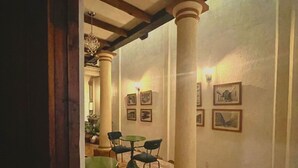 Uma área de refeições com piso xadrez, vigas de madeira e colunas. Há mesas e cadeiras, e luminárias fixadas na parede.