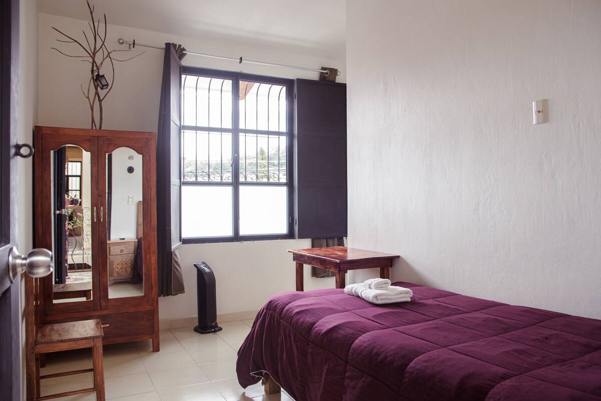 Un dormitorio con cama, armario de madera, espejo, ventana con cortinas y una mesita.