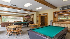 A pool table with surrounding chairs, a dining table, and a bar area with a stone wall and windows. - MY LOSS IS YOUR GAIN!  1 bed Villa at MountainLoft (Gatlinburg)