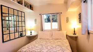 Ein Schlafzimmer mit einem Bett, zwei Nachttischen, einem Fenster mit Vorhängen, einem an der Wand befestigten Spiegel und einem Regal mit verschiedenen Gegenständen.