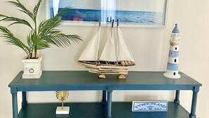 Een donkerblauwe houten console tafel met twee lades, een bloempot, een miniatuur zeilboot, een model van een vuurtoren en twee ingekaderde foto’s van kustlandschappen.