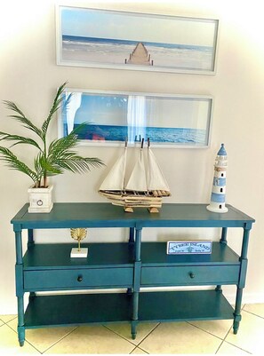 A dark blue wooden console table with two drawers, a potted plant, a miniature sailboat, a lighthouse model, and two framed pictures of coastal scenes.