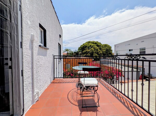 Un balcon avec une table et des chaises, un bâtiment blanc, une clôture en métal noir et un arbre en arrière-plan.
