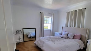 Ein Schlafzimmer mit einem großen Bett, einem Nachttisch, einem Spiegel und einem Fenster mit Vorhängen.
