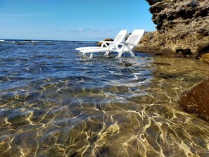 Two white lounge chairs partially submerged in clear shallow water with rocky terrain and a cliff in the background.