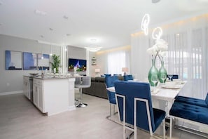 A modern kitchen with a central island, a dining area with blue upholstered chairs, and a living area with a sofa and a TV.