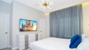Een slaapkamer met een groot bed, een flatscreen televisie aan de muur, een moderne kroonluchter en een groot raam met gordijnen.