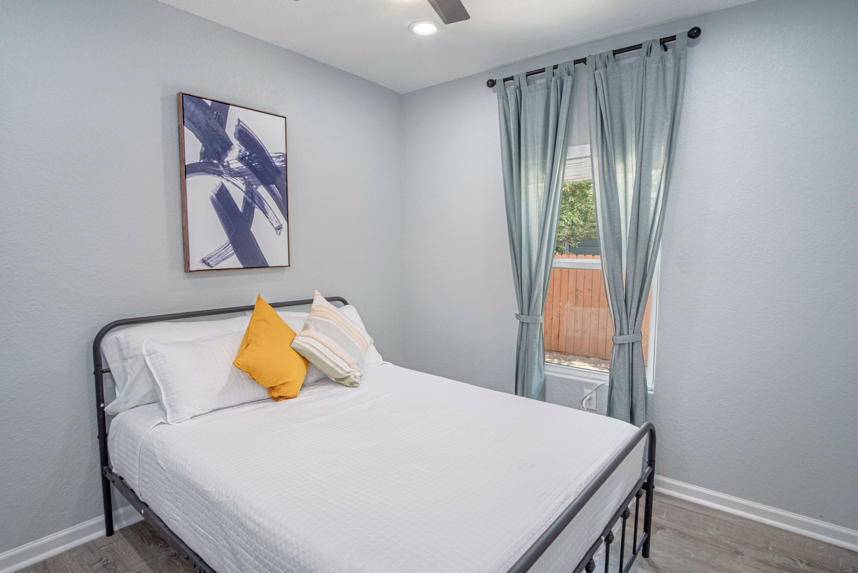 Una cama bien tendida con ropa de cama blanca y una almohada amarilla, un cuadro abstracto enmarcado en la pared y una ventana con cortinas azul claro.