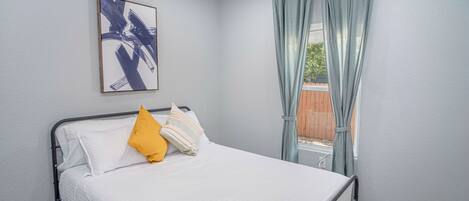 Una cama bien tendida con ropa de cama blanca y una almohada amarilla, un cuadro abstracto enmarcado en la pared y una ventana con cortinas azul claro.