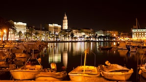 A marina at night with boats docked, a prominent tower, and illuminated buildings along the waterfront.