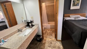 A hotel room with a bathroom featuring a sink, mirror, and shower. There is a bed with a headboard and pillows. - BIGGS NU-VU MOTEL (Biggs Junction)