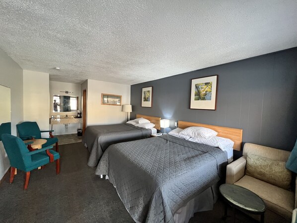 Habitación de hotel con dos camas, una zona de estar con una silla y una mesita, un espejo y obras de arte en las paredes.