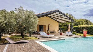 A pool area with a wooden deck, lounge chairs, a covered patio, and a yellow building with a door.