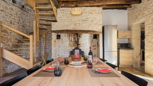 Een rustieke eetkamer met een houten tafel gedekt voor vier personen, een stenen muur, een houten trap en een zichtbare keuken door een open doorgang.