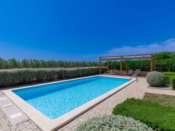 A rectangular swimming pool with a wooden deck and lounge chairs, surrounded by well-maintained hedges and greenery.