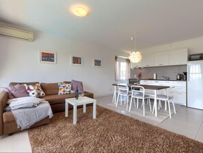 A modern living room with a brown sofa, white coffee table, and a brown shag rug. There is a dining area with white chairs and a kitchen with white cabinets and appliances.