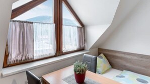 A room with a sloped ceiling, a large window with curtains, a wooden table, a chair, a small potted plant, and a bed with pillows. - Harrachov Central Nest by Interhome (Harrachov)