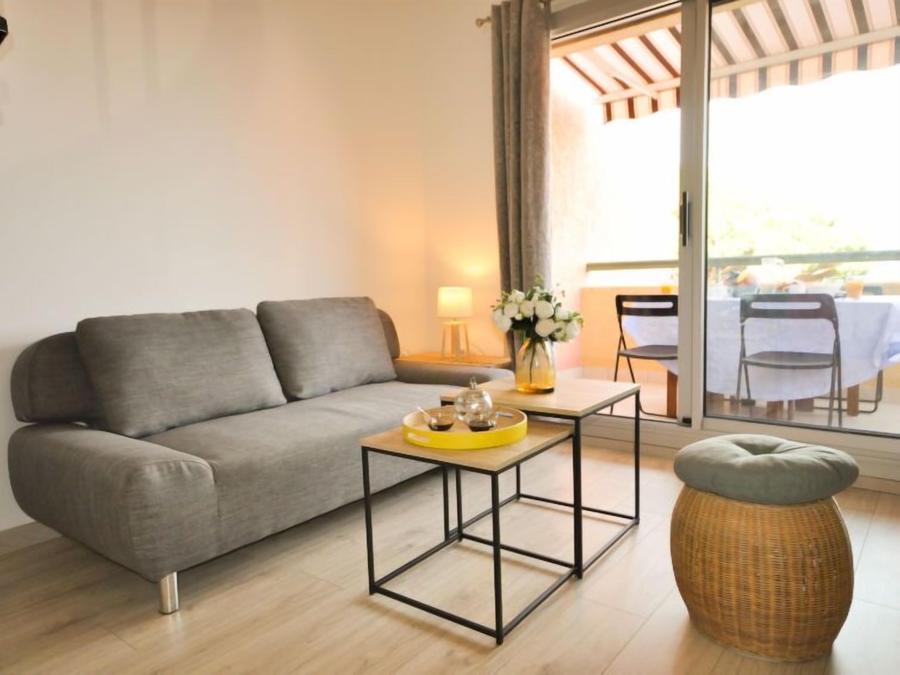 A modern living room with a grey sofa, a small coffee table, a wicker ottoman, and a view of an outdoor dining area through a glass door.