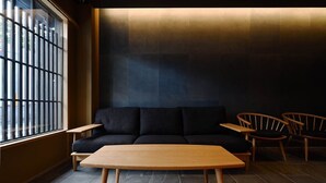 A modern interior with a dark sofa, wooden chairs, a wooden table, and a large window with bars. - FOUR SEASONS INN KYOTO (Kyoto)