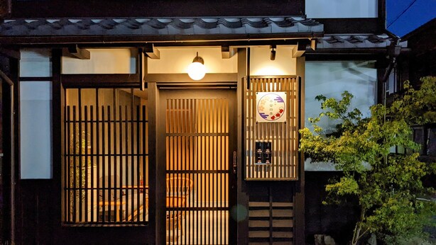 A traditional Japanese building with a wooden gate, a sign with a circular emblem, and a small tree in the foreground. - FOUR SEASONS INN KYOTO (Kyoto)