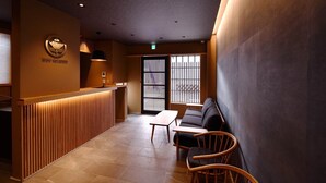 A reception area with a wooden reception desk, a sofa, and two chairs. There is a large window with vertical blinds, and the walls are a warm brown color. - FOUR SEASONS INN KYOTO (Kyoto)